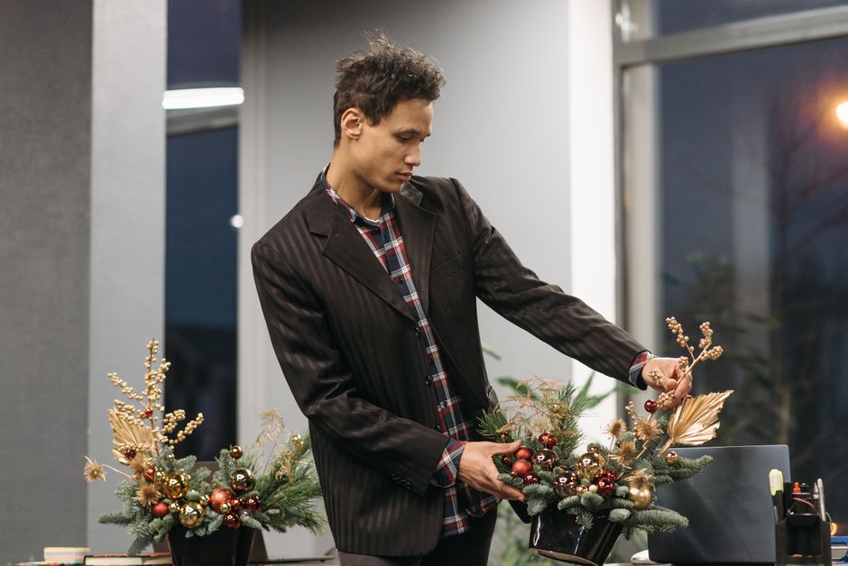 A young man with curly brown hair, wearing a dark pinstripe blazer over a red and black plaid shirt, arranging a festive floral display on a table. The arrangement features an assortment of fresh flow
