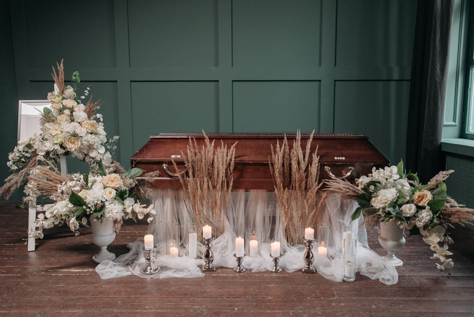 A floral arrangement set on a wooden surface features two large white vases filled with white roses, hydrangeas, and other soft-colored blooms, accented by dried pampas grass and greenery. Tall, slend