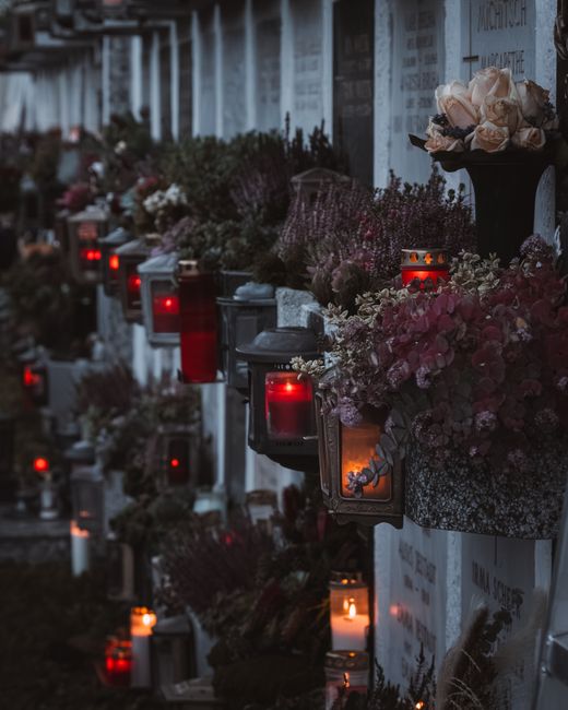 A row of memorial flowers and candles placed on a gravestone wall, featuring soft pink roses, white baby's breath, and purple flowers arranged in a bouquet, with some flowers wrapped in decorative pap