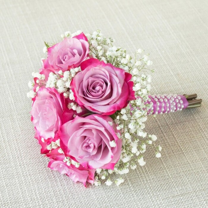 A compact and luminous floral arrangement, this wedding bouquet by Finsbury Flowers speaks to the heart of local North London celebrations. Five vibrant roses take centre stage, each bloom shimmering with a captivating transition: their centres a soft wash of lavender, blending artfully to magenta and rich fuchsia at the edges. Interspersed with sprays of delicate white baby's breath, the bouquet achieves a floating, almost ethereal quality, the tiny flowers catching the light and amplifying the depth of the central blooms. The stems are tightly gathered and finished with a smooth, richly coloured purple satin ribbon-its surface further embellished with spirals of lustrous white pearls that catch and reflect the fine details of the arrangement. Below the elegant wrapping, trimmed stems emerge in natural green-brown hues, anchoring the design. The bouquet is displayed against a pale, textured background-perhaps the kind of classic linen seen in Finsbury's long-standing cafés-helping every hue and detail stand in sharp relief. Whether intended for a modern ceremony at Finsbury Town Hall or a romantic photoshoot in an Islington park, this bouquet blends classic sophistication with local creativity, offering brides a touch of North London's unique charm and poetic spirit.