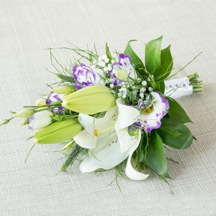 A refined, elongated bouquet is positioned diagonally across a light beige, textured cloth, the neutral background reminiscent of natural linen and giving the entire image a soft, elegant calm often seen in Finsbury townhouse interiors. Dominating the arrangement are several large white Oriental lilies, their petals thick and almost velvety, opening widely to display the warm yellow-orange stamens and delicate speckling near the flower's throat. Some of the lilies are fully unfurled, while others are just beginning to open, creating a sense of time unfolding. Around them sit sleek, bright green lily buds, still closed and elongated, offering a promise of continued bloom long after the bouquet is delivered. Threaded through the lilies are clusters of Eustoma, or lisianthus, their petals forming gently frilled cups that shift from creamy white at the base to bold purple at the edges, with tones ranging from gentle lavender to deep violet, like brushstrokes of colour. Fine sprigs of baby's breath weave in and out, their countless tiny white blossoms giving a light, misty effect that softens the more dramatic blooms. A rich base of foliage supports the flowers: broad, glossy dark leaves provide a strong, luxurious foundation, while finer, wispy greenery and a few slender, upright stems introduce texture and lively movement, as though catching a breeze drifting across Finsbury's open squares. At one end, the stems are neatly aligned and bound with a smooth white ribbon, accented with delicate, pearl-like beads in an even line that hints at bridal detail and craftsmanship. The lighting is gentle and diffused, free of harsh shadows, making the whites, greens, and purples appear fresh and luminous. This bouquet feels perfectly suited for a wedding near Finsbury, an anniversary dinner at home, or a thoughtful, hand-delivered gift from Finsbury Flowers to someone special in the neighbourhood.