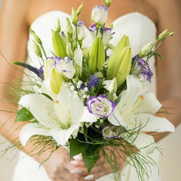 Embodying refined beauty in Finsbury style, this close-up showcases a stunning hand-tied bridal bouquet, radiant in the bride's gentle grasp. Several large, pure white Oriental lilies stand tall at the bouquet's centre, some fully open and displaying their intricate spotted petals and sun-bright stamens, while others are in soft transition from bud to bloom, bearing subtle green highlights. Interspersed throughout is a scattering of lisianthus-white, ruffled, and edged delicately in lavender-evoking the artisanal spirit of the area's florists and the relaxed elegance of Roseberry Avenue's social gardens. The rich purple spikes of Veronica or Muscari emerge boldly, imparting striking depth and echoing the lively market stalls of Exmouth Market. Wisps of classic baby's breath and finely textured light green asparagus fern surround the florals, softening the arrangement and lending it an unstudied air of natural beauty. Broad, glossy leaves and unopened lily buds create depth and balance, while the bride's hands, manicure immaculate, lovingly cradle the arrangement. The softly blurred white dress and bare shoulder in the background place the focus firmly on these exceptional blooms. Crafted for ceremonies at Myddelton Square or celebrations near Mount Pleasant, this Finsbury Flowers bouquet embodies timeless romance for modern urban weddings.