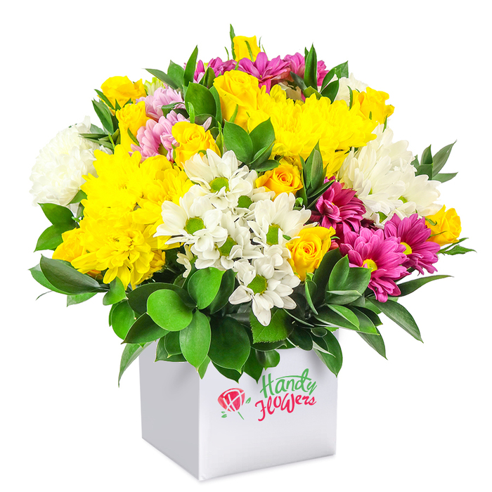 A joyful, dome-shaped floral arrangement is presented in a simple white square box, photographed against a pure white background that allows every colour to stand out clearly. The bouquet is strikingly full and rounded, with a dense mix of blossoms that creates a sense of rich, layered texture. At its heart are generous yellow chrysanthemums, their many fine petals forming soft, cushion-like blooms that catch the light, alongside clusters of yellow spray roses in varying stages-from neat buds to partially open flowers that reveal their spiralled centres. Between these golden highlights nestle cool white flowers: small daisy-like chrysanthemums with bright green eyes, and larger, fluffy white chrysanthemums that lend softness and volume to the arrangement. Running through this field of yellow and white, rich fuchsia and magenta chrysanthemums add visual drama, their saturated tones giving the bouquet a contemporary flair that would look at home in a Finsbury townhouse or a stylish flat near Clerkenwell. Delicate pink and extra yellow rosebuds are dotted throughout, offering small, intimate points of interest that reward a closer look. The entire composition is wrapped in a collar of deep green, glossy foliage that peeks between the blooms and frames the base, adding contrast and emphasising the fresh, just-arrived-from-the-market feel. The white cube box functions as both vase and gift packaging, with its clean lines and smooth surfaces underscoring the modern aesthetic favoured by many local Finsbury florists. This arrangement feels perfect for marking happy moments-a birthday breakfast near Finsbury Park, a simple thank-you, or a spontaneous gesture to brighten a friend's day with same-day delivery from Finsbury Flowers across the area.