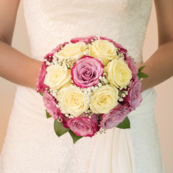 In the heart of Finsbury, a vibrant, close-up scene unfolds: a bride, her midsection framed against the light of a softly blurred background, embraces a round bouquet that gleams with contemporary elegance and traditional floral flair. The composition draws the eye-dozens of tightly clustered roses create a tapestry of creamy yellow and vivid fuchsia, accented by hints of lavender-mauve, echoing the dynamic colours seen in Finsbury's Green spaces. Sprigs of baby's breath weave whispery threads throughout the bouquet, providing lightness and softness reminiscent of clouds drifting above Rosebery Avenue. A scattering of deep, glossy green leaves radiates from the base, grounding the ensemble and connecting it to the local parks. The wedding dress, white as freshly fallen snow, is characterized by exquisite lace motifs across the bodice, while the skirt tumbles in cascades of smooth fabric, all illuminated by an aura of gentle daylight. The bride's arms arch gracefully around the bouquet, capturing a poised and affectionate embrace. This photograph captures not just a beautiful arrangement but the emotion of a Finsbury celebration: the freshness, the anticipation, and the artistry that Finsbury Flowers brings to weddings in this beloved London neighbourhood.