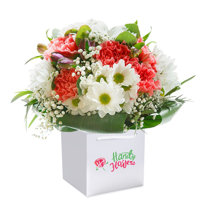 A vibrant, thoughtfully composed bouquet radiates brightness from within a fresh white square gift bag, emblematic of Finsbury's modern elegance and friendliness. Ruffled coral and soft peach carnations form the heart of the design, their full forms and rich textures capturing the easy confidence of the neighbourhood's gardens. White daisy-like chrysanthemums with their green-centred eyes appear in playful clusters, their purity highlighted by interwoven sprays of delicate baby's breath that seem to hover like early morning mist above Finsbury's secluded garden squares. Hidden amongst the open petals, closed buds tinged with purple and pink lend an air of intrigue, reminiscent of the hidden corners of Finsbury you stumble upon by chance on a Sunday stroll. Framing the entire arrangement are broad leaves, slick and deep green, poised in an elegant oval that grounds the cheerful palette above. The bouquet perches in a white bag, complete with soft ribbon handles that invite gifting or easy delivery down the lanes of Pentonville or towards the bustle of King's Cross. Under bright but gentle lighting, the colours sing with freshness and hope-making this ensemble an ideal present for birthdays, milestones, or a spontaneous gesture of care, thanks to the artistry and local pride that characterise Finsbury Flowers.