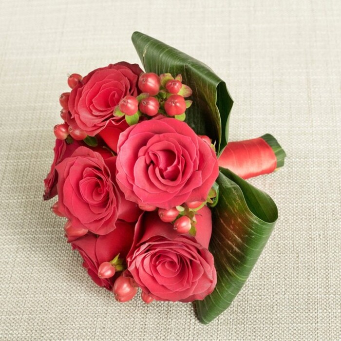 A lush cluster of deep red roses and berries forms a compact, sculpted bouquet displayed on a soft, light beige fabric that looks like finely woven linen, its gentle texture visible beneath the blooms. The arrangement is captured from a slightly raised viewpoint, so the viewer can appreciate the rounded silhouette and layered composition. At the centre, about six large roses in saturated shades of crimson and rich scarlet dominate the scene, their velvety petals spiralling outward from tight hearts into open, cup-shaped blooms that seem almost touchably soft. Nestled among the roses, numerous small Hypericum berries in red and reddish-pink tones stand out, glossy and bead-like, each cluster held by delicate green stems that slip between the petals, adding festive detail and a sense of abundance. On one side, two prominent dark green leaves with clearly defined veins are gracefully curved and wrapped around the bouquet, functioning like a natural frame that holds the flowers in a neat, modern form. Additional deep green foliage fills in the base, enhancing the colour contrast and giving the bouquet a grounded, organic feel. Around the stems, a smooth red satin or silk ribbon is neatly tied and tightly wrapped, its rich sheen harmonising with the roses and hinting at careful, professional craftsmanship. The neutral, softly lit background lets the reds and greens glow, as though the bouquet has been placed on a prepared table for a celebration in a Finsbury loft near Old Street. This arrangement feels ideal for anniversaries, intimate weddings, or Valentine's Day deliveries across the EC1 area, its classic palette conveying romance, devotion, and a refined sense of London style.