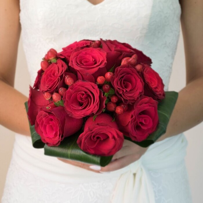 In this evocative waist-up portrait, a bride-her form draped in the distinctive lace and soft fabric of a Finsbury wedding gown-holds an exquisite bouquet crafted with local expertise. The primary attraction is a tightly clustered and rounded assembly of plush, ruby red roses, each bloom at varying stages of unfurl, their velvety petals spiraling outward with depth and vibrancy. Between the roses, vivid red hypericum berries are interspersed, providing a textural lift and a jewel-like brightness akin to the vibrant displays at Whitecross Street Market. The bouquet is wrapped with expertly folded broad leaves, dark green and glossy, reminiscent of the healthy botanical borders seen in Finsbury's parks. The bride's hands, neatly manicured and gentle, support this luxurious bouquet, set off perfectly against her gown's intricate lacework, whose details echo the local area's historic charm. The overall atmosphere is one of warmth, depth, and quiet celebration, brought to life by soft, natural light and the loving attention of Finsbury florists. This classic arrangement celebrates passion and sophistication, making it an ideal centrepiece for ceremonies or as inspiration for local bridal features.