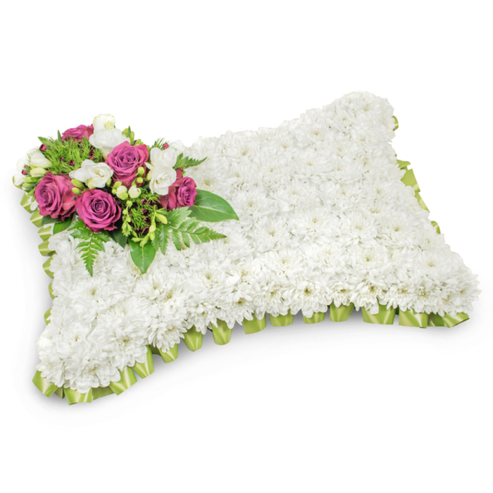 A rectangular memorial cushion, meticulously covered in a dense carpet of small white chrysanthemums, reads at first glance like a plush, comforting pillow. Each tiny bloom is placed to produce a uniform texture and a serene, almost fabric-like surface that evokes peace, purity and the idea of eternal rest. Nestled on the top-left of the form is an intentional, graceful spray of deep magenta and fuchsia roses - their saturated tones and layered petals forming a focal blossom cluster that contrasts beautifully with the surrounding white. Scattered among those roses are small white freesias or similar delicate buds whose scent is light and clean, and whose shape introduces a delicate punctuation to the composition. Lush greenery, including fine fern fronds and glossy leaves, fans beneath and around the accent cluster to provide depth, movement and a restorative green base. The perimeter is finished with a pleated light green satin ribbon, looped or tucked to create a refined border that just peeks out from the flowers and lends an understated ceremonial edge. Against a bright white backdrop and a soft shadow below, the pillow's three-dimensional form is quietly dignified. This traditional sympathy arrangement - designed for funeral services or graveside placement - offers a tender visual metaphor for rest and remembrance, and is the kind of thoughtful tribute families in Finsbury, near Clerkenwell and the Barbican, often choose to honour a loved one.