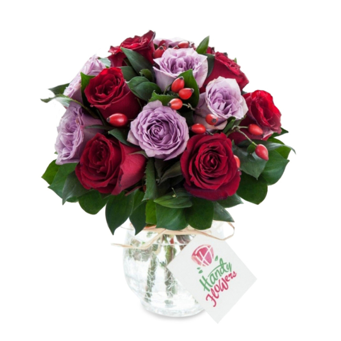 An elegant, densely arranged bouquet of roses combining deep burgundy and crimson tones with pale lavender and lilac blooms, presented as a compact, rounded spray that feels both sumptuous and intimate. Each red rose shows layered, velveteen petals, some with inner petals still gently unfurling, while the purple roses bring a soft, powdery hue that tempers the intensity of the reds; together they create a rich, romantic contrast that reads as sophisticated and heartfelt. Lush, dark green foliage-likely Ruscus-sits throughout the design, its smooth leaves offering structure and a cool matte sheen beneath the flowers. Bright red-orange rose hips or berry clusters are artfully tucked into crevices between blooms, adding a rustic, tactile accent and a pop of autumnal colour that complements the classic rose palette. The flowers are arranged in a vintage-style mercury glass vase: rounded at the base, slightly tapered at the top, its antiqued silvered surface mottled and reflective, catching glints of the flower colours. A natural raffia tie encircles the vase's neck and a small white rectangular gift tag, featuring a simple rose illustration and soft handwritten text, hangs forward to suggest a personal message. Photographed on a clean white background, the bouquet feels freshly gathered and ready for delivery-an arrangement I might prepare for a client picking up from Exmouth Market or requesting same-day delivery across Finsbury. Crafted with neighbourhood care by local hands, it balances romance with a grounded, market-fresh sensibility that clients expect from Finsbury Flowers.