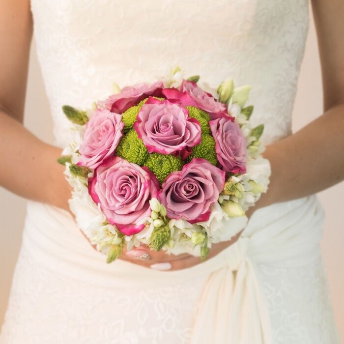 Positioned elegantly against an intricate off-white wedding dress, a bride in Finsbury gently supports a bouquet designed with both tradition and modern flair by a local florist. At the bouquet's center stand several lavender-pink roses-each bloom opening fully, its color softly shifting to a vibrant fuchsia at the edges, mirroring the passion and sophistication associated with Finsbury's creative community. Nestled among them, lime-green pom-pom chrysanthemums provide a vivacious punch of color and a playful counterpoint to the softness of the roses, adding a feathery texture to the tightly rounded bouquet. Delicate white flowers, possibly sweet scented freesia or clusters of small spray roses, create a dreamy halo along the edge, their subtle fragrance filling the air. The piece is finished with touches of green buds and slim leaves, offering a hint of wild but artful abundance. The bride's gently curved fingers, nails decorated in serene blush and a single fingertip sparkled for subtle festivity, are illuminated by radiant natural light-a scene easily imagined just steps from Finsbury's Clerkenwell Green or Rosebery Avenue. This bouquet embodies the seamless union of classic romance and vibrant urban personality, ideal for couples celebrating their love within the heart of North London.
