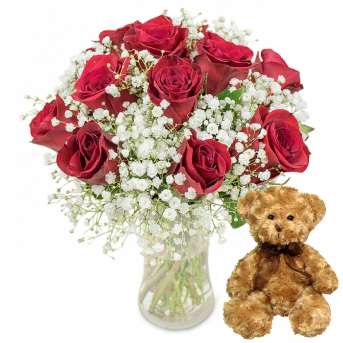 Captured against a pristine white studio background, this image portrays a beautifully balanced floral and gift combination, the kind of arrangement a florist at Finsbury Flowers might prepare for a surprise delivery near Angel or Old Street. On the left sits a clear glass vase, modest in design yet elegant in its simplicity, half-filled with water that magnifies and distorts the bundle of stems within. Rising from the vase, about twelve red roses are arranged in a carefully shaped, generously rounded cluster. The roses display velvety petals in lush layers of crimson, with some blooms opening wide to reveal intricate spirals and others held slightly tighter, their centres still curling inward in a promise of further unfolding. Interspersed among the roses is a profusion of white baby's breath (Gypsophila), its countless tiny blossoms gathered along slender stems. These delicate flowers create a fine, airy mist around the more substantial rose heads, softening the intense red and adding a gentle, almost ethereal quality to the composition. Glimpses of dark green foliage and the clear lines of the stems lend the arrangement a fresh, just-cut-from-the-market feeling. To the right of the vase rests a medium-sized teddy bear, seated in a relaxed, upright position. The bear's fur is a light, warm brown, appearing plush and invitingly soft, with rounded paws resting calmly in front. Its face is endearing, with small, shiny dark button eyes, a petite embroidered nose, and a faint stitched smile that hints at quiet friendliness. A narrow dark brown satin-style ribbon is tied neatly in a bow around its neck, completing the bear's classic look. Together, the vibrant roses, airy gypsophila, and cuddly bear evoke themes of love, tenderness, and celebration, making this visual ideal for illustrating romantic flower and gift delivery options in Finsbury and nearby central London districts for occasions such as anniversaries, Valentine's surprises, or special birthday moments.