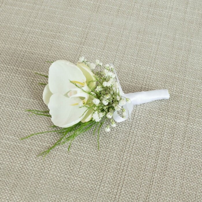 A refined white orchid boutonniere is captured resting on a pale beige, woven fabric backdrop, whose subtle linen-like texture brings to mind the table runners and chair sashes often seen at stylish wedding venues around Finsbury and along Rosebery Avenue. At the centre of the composition sits a single, immaculate Phalaenopsis orchid bloom, its petals broad, rounded, and velvety, unfolding in layers of pure white. Toward the throat of the orchid, barely-there touches of soft yellow and creamy tones add depth, as though a beam of sunlight has settled gently in its heart. Surrounding this central flower is a soft ring of baby's breath, each Gypsophila stem branching into tiny, snowflake-like blossoms, so small and numerous that they create a misty, cloud-like aura around the orchid. Threaded through the baby's breath are slim, feathery strands of fresh green foliage, reminiscent of delicate ferns or similarly light greenery, introducing a lively contrast while keeping the overall tone airy and romantic. The stems are skillfully aligned and bound with a smooth, matte white satin ribbon, wrapped snugly and finished in a simple, elegant bow that suggests careful craftsmanship and attention to detail. The lighting is gentle and even, casting minimal shadow, which allows the crisp whites, soft creams, and bright greens to stand out clearly against the neutral background. The overall mood is one of timeless simplicity and modern grace, perfect for a groom, best man, or wedding guest attending a ceremony at a local Finsbury church or registry office. It feels like the kind of bespoke lapel flower a couple would choose when working closely with a trusted neighbourhood florist to create a look that is both classic and quietly contemporary.