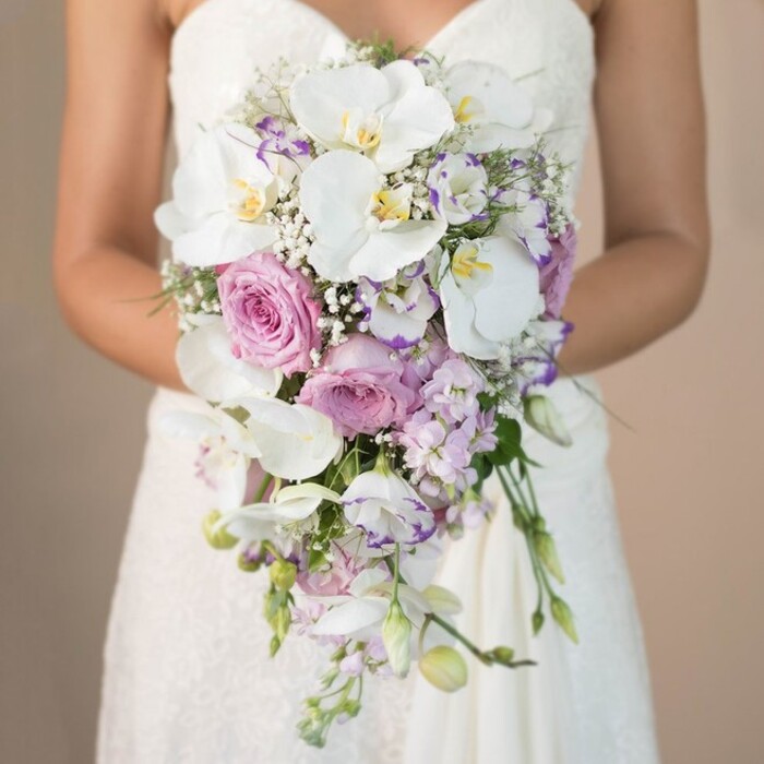 This close-up, portrait-style photograph captures the essence of a Finsbury bride: poised, elegant, and holding a bouquet as unforgettable as the city's blend of tradition and modernity. Framed by a muted, blurred beige background, the focus rests entirely on the bride's hands, gently clasping a flowing, tear-shaped bridal bouquet. At its heart are sumptuous white phalaenopsis orchids, each with satiny petals and gold-tipped throats, surrounded by dreamy, billet-doux pink garden roses and exquisite lavender stock. Threaded among them are clusters of white lisianthus, their edges sometimes blushed with purple, and ethereal touches of baby's breath for an airy, sentimental feel. Sparse sprigs of green foliage lend freshness, echoing the green spaces like those surrounding Finsbury Square or beneath the trees on Myddelton Passage. The bride's strapless lace gown, just visible, blends seamlessly into the scene, evoking the romance of a city wedding in Clerkenwell or near Exmouth Market. Every element in the arrangement, crafted by a team of expert florists at Finsbury Flowers, blends seasonality and beauty into a bouquet that exudes elegance, softness, and a touch of North London sophistication-ideal for couples whose love stories intertwine with the local community.