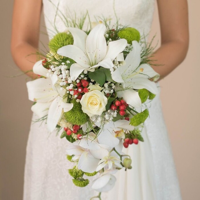 At a civil ceremony venue near Finsbury's elegant gardens, a close-up image reveals a bride in a lace-detailed ivory gown holding a gorgeously cascading bridal bouquet. The composition is a study in harmony - luminous white oriental lilies, each petal bearing modest rust-hued markings, radiate graceful sophistication at the bouquet's crown. The bouquet's heart features a tightly wound, cream-white rose, a timeless emblem of a love newly pledged. Interspersed amidst these blooms are lively bursts of lime-green chrysanthemum, their contemporary colour drawing inspiration from Clerkenwell Green's lush gardens. Tiny hypericum berries, their bright red hue reminiscent of festive local celebrations, cluster between the lilies and mums, adding lively warmth and seasonal cheer. Throughout the bouquet, delicate clouds of baby's breath weave a gentle lightness, their fine texture softening the scene like mist along Finsbury's side streets. Drawing the arrangement together, elongated grass sprigs and rich foliage create a natural, flowing edge, while elegant white whorls of Phalaenopsis orchid drift gracefully downwards, giving the bouquet its dramatic waterfall shape. The bride's hands, lightly clasping satin ribbon at the stems, anchor the burst of blooms against the subtle backdrop of intricate lace. This superb, locally inspired bouquet by Finsbury Flowers unites tradition, style, and a fresh palette - a perfect reflection of both the neighbourhood's history and the contemporary romance found in today's weddings.