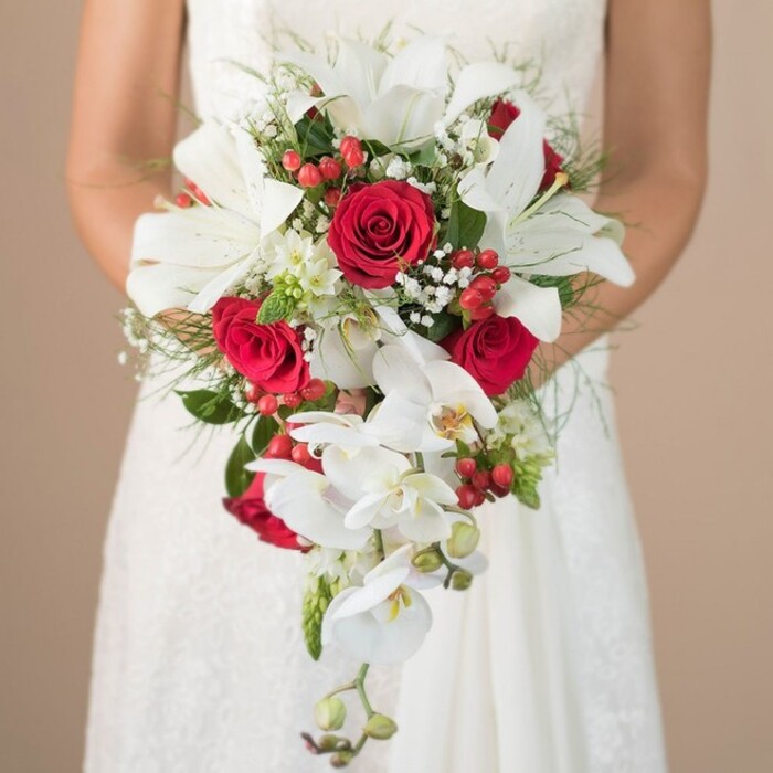 Bathed in soft daylight, this stunning bridal bouquet takes centre stage as a bride stands poised in her elegant white gown, just steps from the historic heart of Finsbury. The arrangement is a feast of colour and form, designed in a cascading teardrop style that evokes sophistication and grace often seen at local Islington celebrations. At its core are striking white lilies, their petals fanned wide to reveal sculptural yellow stamens, while trailing white orchids flow seamlessly downwards, echoing the fluid artistry of Finsbury florists. Interlaced amid these centrepieces, lush red roses unfold with a timeless elegance, each velvety bloom catching the light alongside clusters of brilliant red hypericum berries. Feather-soft clouds of baby's breath drift through the bouquet, offset by fine green asparagus fern and deeper green leaves reminiscent of the verdant corners in Finsbury's parks. Spiky foliage elements peek among the blooms, adding intrigue and wildness to the refined design. The bride's lightly tanned arms provide gentle support, her lace-trimmed gown catching delicate highlights. Set against a neutral, blurred backdrop, this floral creation encapsulates passionate romance and local craftsmanship, offering inspiration for any London couple planning a memorable Finsbury wedding.