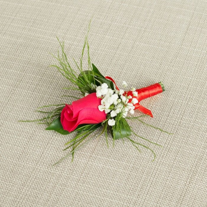 A meticulously arranged boutonnière is displayed on a pale, linen-like fabric, the soft beige background allowing every colour and texture to stand out with clarity. At the centre is a single rosebud in a vivid fuchsia or deep magenta tone, its petals tightly wound and almost velvety to the eye, suggesting a rose at the peak of freshness, as if it had just been delivered from a local Finsbury florist. Tiny florets of snow-white baby's breath cluster snugly around the base of the rose, forming a gentle, airy halo that balances the intensity of the magenta. Behind and around the blooms, a selection of greenery has been chosen with care: broader, darker green leaves provide structure and weight, while slender, wispy, grass-like sprigs arc outwards, adding a natural, slightly wild elegance reminiscent of the planting in nearby London garden squares. The stems of the entire arrangement are drawn together and wrapped in a vibrant red satin ribbon, wound smoothly and tightly, with a neat little bow or knot at the base. The ribbon's glossy surface catches the light and echoes the warmth of the rose, while also offering a secure grip for pinning it to a lapel. The boutonnière is angled slightly upwards, creating a sense of movement and making it easy to imagine it resting on the jacket of a groom, best man, or prom date heading out through Finsbury's streets. The photograph is well lit, revealing every nuance in the rose petals and the fine texture of the baby's breath, conveying romance, attention to detail, and a timeless sense of formal style.