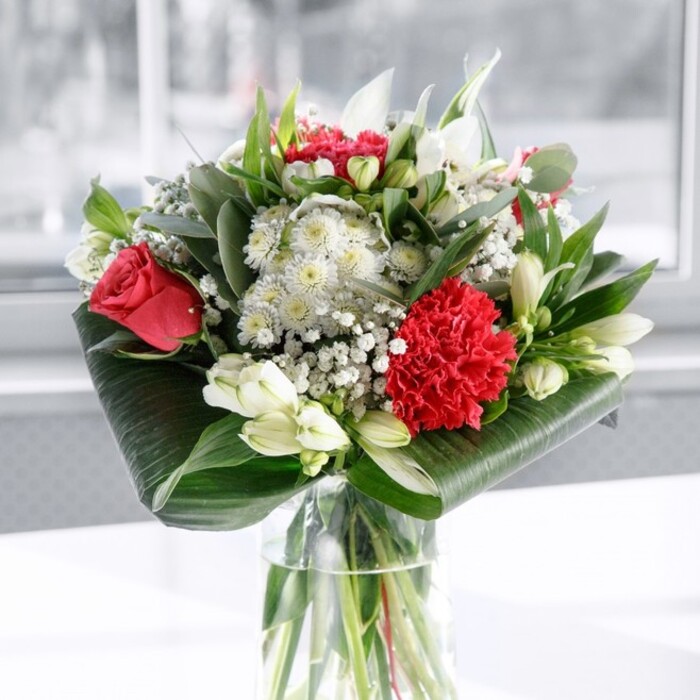 A lush bouquet in classic red, white, and green stands tall in a clear cylindrical glass vase, its stems neatly aligned and visible beneath the water, suggesting the meticulous work of a seasoned florist from the Finsbury area. At the heart of the arrangement, velvety deep red roses open in soft spirals, their petals just beginning to unfurl, while a large crimson carnation with densely ruffled edges anchors the front, giving the arrangement a sense of movement and texture. Surrounding these rich red blooms are elegant white flowers: graceful Alstroemeria with slim, slightly flared petals marked by fine internal speckles, alongside several tightly closed white lily buds whose smooth, tapered forms hint at the fuller blossoms to come. Wisps of airy gypsophila weave through the bouquet, their tiny white florets resembling a dusting of light across the composition, softening the bold colours and adding a romantic, almost ethereal layer. The flowers are cradled and framed by abundant greenery, from broad, glossy dark green leaves wrapped and curled artfully at the base to upright, pointed foliage and eucalyptus-like stems that add height, variation, and a subtle silvery undertone. The bouquet is set on a pristine white surface, with a softly blurred background washed in gentle brightness, evoking the feel of natural daylight streaming through a London window near Finsbury's bustling streets or the quieter corners by local parks. The even lighting draws out the richness of each petal and the shine of each leaf, creating a balanced, harmonious look. This arrangement would make a beautiful centrepiece for a family celebration in a Finsbury townhouse, an anniversary dinner in a local restaurant, or as a sophisticated gift delivered to an office, conveying affection, admiration, and an air of timeless romance.