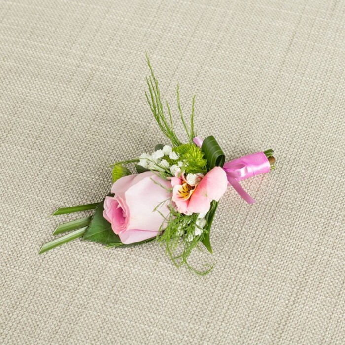 Against a softly woven, creamy linen background that brings to mind Finsbury's urban tranquillity, an expertly composed boutonniere is thoughtfully arranged. The centerpiece, a pastel pink rosebud, boasts petals closely wrapped in a graceful spiral, transitioning from a dusky heart to pale edges, echoing the refined gardens of Myddelton Square. Next to it, a delicate alstroemeria with faint peach and yellow tones opens wide, its subtle markings evoking the fine detailing often found in historic Finsbury residences. Sprigs of baby's breath introduce gentle, cloudlike lightness, interspersed with lively green button mums resembling freshly polished park lawns. Outreaching blades of deep green grass add elegant height, balanced by bold, broad leaves folded and cupped at the boutonniere's base; one larger leaf securely sheaths the stems. The entire design is bound together by a satiny fuchsia ribbon, tied in a small, luminous bow that imparts vibrancy and celebration. The neutral fabric setting highlights every floral nuance, from soft hues to crisp textures, while the overall arrangement glows softly under ambient lighting-an inviting accent ideal for special events, weddings, or proms in Finsbury or nearby Islington. This boutonniere, meticulously crafted by Finsbury Flowers, is a celebration of local artistry and heartfelt emotion.