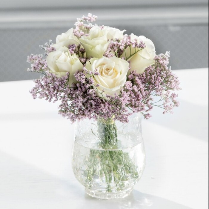 At the centre of the composition, a meticulously arranged, round bouquet of creamy white roses and lilac statice stands in a tall, cylindrical clear glass vase, placed on a pristine white tabletop that reflects the soft daylight. The roses, about a dozen in all, present as full, perfectly formed blooms, their petals a gentle shade somewhere between ivory and warm cream. Each rose is captured in that fleeting moment when the outer petals just begin to curl open, revealing a layered spiral of softness inside, with slight variations in tone that add natural depth. Interwoven among these classic blooms are generous clusters of fine-stemmed statice, their tiny, lavender-purple flowers creating a gauzy halo around and between the roses. The statice adds not only colour but intricate texture, its papery, almost crinkled florets catching light differently from the smooth rose petals, giving the entire bouquet a lively yet delicate structure. Through the clear glass of the vase, the green stems are visible in tidy vertical lines, immersed in fresh, transparent water that hints at attentive, professional conditioning-exactly the standard a local shop like Finsbury Flowers would insist upon before delivering to homes and offices along Farringdon Road or around Old Street. The bright, uncluttered white surface beneath suggests a contemporary interior, perhaps a minimalist flat near Finsbury Square or a calm meeting room looking out toward the City. The background is softly out of focus, suffused with pale natural light that seems to stream from a nearby window. This gentle illumination makes the roses appear almost luminous, while faint, diffuse shadows on the table enhance the sense of depth. The atmosphere conveyed is one of quiet refinement and emotional restraint, making this arrangement ideal for a wedding morning, an elegant anniversary surprise, or a sympathetic gesture that brings a touch of polished serenity to any Finsbury setting.