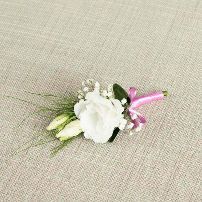 An elegant wedding boutonnière lies at a gentle angle on a light beige, textured fabric that resembles finely woven linen, the natural fibres creating a subtle pattern under soft, even lighting. At its centre, a fully bloomed white lisianthus steals the eye, its ruffled petals opening wide in concentric layers that form a soft, rounded cup. The flower's tone is a pure, creamy white, with delicate petal edges that look almost like fine tissue, catching the light and giving the bloom a luminous, graceful presence. Tucked to the lower left side are two elongated lisianthus buds, still closed, their smooth, tapered forms shaded in gentle gradients of pale green and cream. They sit close to the main flower like quiet companions, suggesting the promise of future blossoms and the unfolding of a wedding day somewhere around Finsbury's leafy streets and garden squares. Interspersed among the central bloom and buds are airy sprigs of gypsophila, or baby's breath, each stem dotted with tiny white flowers that create a cloud-like softness around the boutonnière. Slender, dark green, grass-like leaves sweep outward to the left, adding a sense of dynamic movement and a light, feathery texture, while a single broader, deeper green leaf rests behind the lisianthus, offering contrast and structure. The stems are gathered neatly together and tightly wrapped at the base with a vivid fuchsia satin ribbon, tied into a small, cheerful bow facing the lower right. The satin catches the light with a gentle sheen, and the energetic pink hue pops dramatically against the neutral fabric and white petals, giving the design a distinctly modern, celebratory character. Photographed from an overhead, slightly elevated angle, this boutonnière feels fresh, romantic, and perfectly suited for a groom's lapel at a local ceremony in Finsbury or a reception overlooking nearby Clerkenwell and Angel.