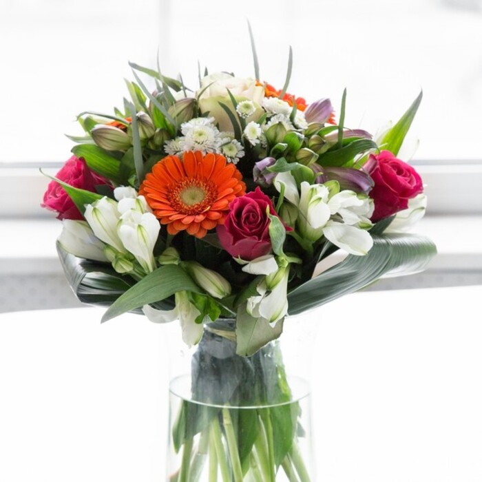 This bouquet, a masterful testament to the artistry of Finsbury florists, weaves together colour, structure, and emotion in a sublime symphony. Soft peach and creamy gerberas form a gentle ring around fiery orange blooms, echoing the warm glow of late afternoons near Amwell Street. The bright, fuchsia-pink roses add depth and richness, introducing a sense of romance reminiscent of walks along the Regent's Canal. Clusters of white alstroemeria and tiny spray roses dot the bouquet, their creamy petals gleaming with freshness and highlighting the care poured into every detail. Broad aspidistra leaves, wrapped decisively around the base, create a bold frame, much like the established greenery in Finsbury's pocket parks offering sanctuary to the district's busy pace. Smaller, pointed leaves contribute lively structure, filling the arrangement with vibrancy and a garden-inspired feel. In its slender glass vase, the display feels modern yet timeless, illuminated by soft light that accentuates every colour and texture. Subtle scents of rose, lily, and verdant foliage linger, conjuring memories of Finsbury's springtime markets. This bouquet, designed for joyful gatherings, thoughtful thank-yous, or celebrating a new chapter, radiates optimism and connection - a floral embrace rooted in the heart of Finsbury.