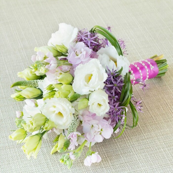 Resting diagonally on a neutral, lightly textured linen-evocative of a Finsbury florist's workshop-this bouquet radiates elegance and lively charm. Creamy white Eustoma (Lisianthus) are the stars, their ruffled, romantic petals unfolding gently around soft yellow-green hearts, speaking of spring's freshness. Interwoven among the white blooms are elongated buds and stems in pale lime and crisp white, capturing the theme of promise carried by London's urban parks. Soft lavender-pink stocks cluster delicately amidst the arrangement, their pastel hues blending seamlessly with the centerpiece while adding a sweet floral scent, much like the early blooms in Finsbury's green squares. For bold contrast, spiky deep purple alliums surge upward, their starry sphered florets forming architectural islands within the softer mass-a playful echo of Finsbury's artistic vibrancy. Fresh green filler elements and gently looping blades of bright foliage add structure and a sense of natural rhythm, imparting movement and depth. The stems are elegantly wrapped in vivid fuchsia satin ribbon and finished with a single strand of gleaming white pearls, a nod to the sophistication expected at Finsbury weddings and festivities. This sophisticated bouquet-ideal for contemporary brides or special gatherings from Rosebery Avenue to Myddelton Square-harmoniously balances romance, color, and an unmistakable local touch.