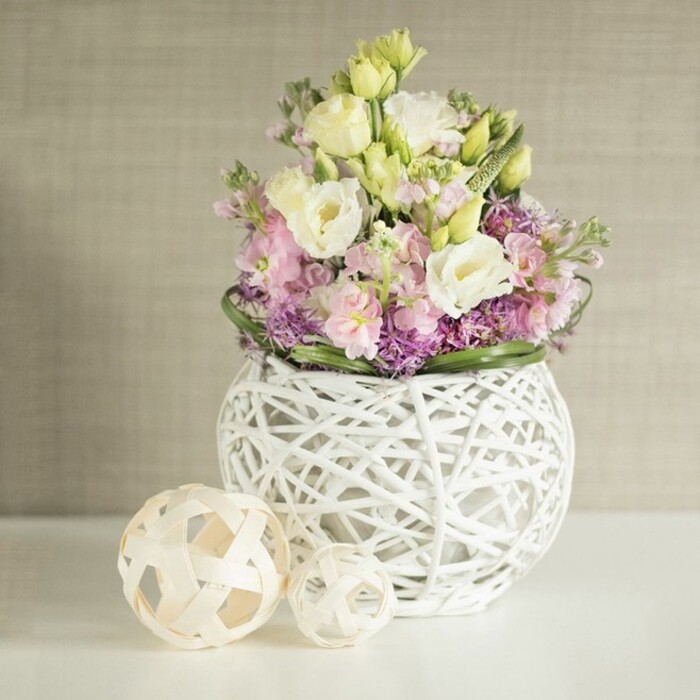A meticulously designed floral centrepiece echoes the welcoming vibrance of Finsbury, displayed on a pristine white table beneath gentle, natural daylight. The central vase-a modern sphere woven from pale, painted rattan-captures the hand-crafted essence found in the area's boutique shops and artisan workshops. Overflowing from its open lattice is a bountiful bouquet of creamy white lisianthus mixed with pale yellow blooms, their signature ruffled petals inviting touch and admiration. Rising amidst these, elegant pink stock flowers stand tall, their muted sweetness and vertical lines reminiscent of Finsbury's formal garden borders. A base of rich purple filler-possibly allium or aster-layering the bottom reinforces the arrangement's opulent texture and lends an air of quiet sophistication. Surrounding the florals is a defined ring of glossy green foliage, much like the manicured plantings found in Myddelton Square. To one side, two decorative spheres made from light wood or rattan mirror the vase's intricate design, their open-work construction creating engaging shadows in the ambient light. The entire arrangement is framed by a soft, linen-textured beige wall, its subtle warmth offsetting the cool tones and fostering a tranquil, elegant atmosphere. This centrepiece embodies the spirit of a Finsbury wedding table, blending local artistry, modern sensibility, and the timeless freshness of expertly curated flowers-available for bespoke arrangements across the neighbourhood by Finsbury Flowers.