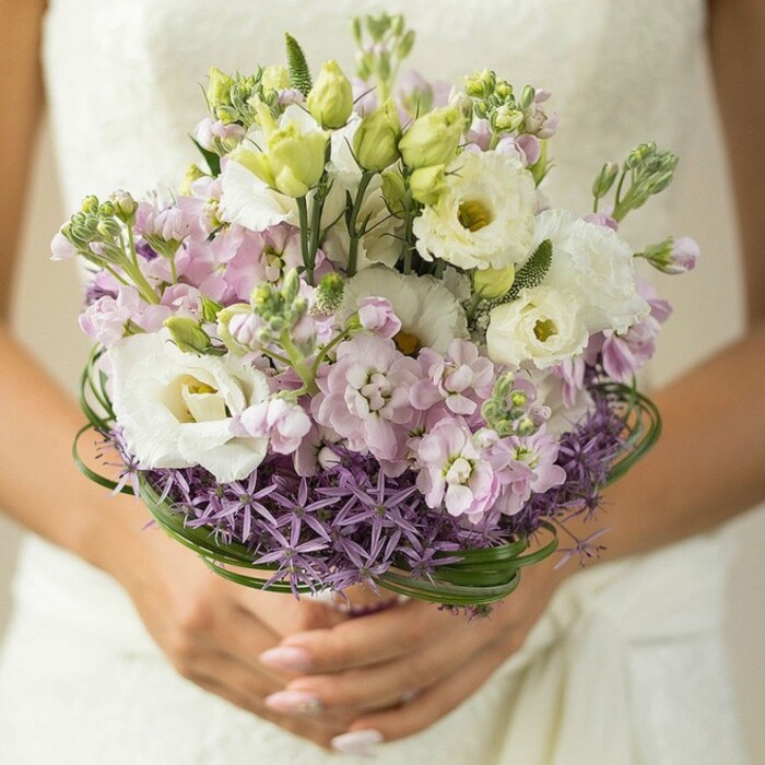A captivating, softly focused image shows a sophisticated wedding bouquet, nestled lovingly in the hands of someone likely adorned in a white or ivory bridal gown. The lower third of the photo reveals perfectly manicured nails in a subtle nude or blush shade, supporting a bouquet of abundant springtime charm. Dominating the arrangement, multiple white Lisianthus blossoms display ruffled, soft petals and pale yellow-green hearts, exuding purity and opulence reflective of Finsbury's refined taste. Interwoven among these are slender, unopened green buds and frothy mounds of lilac and pale pink stock flowers, adding texture, scent, and a gentle transition of hues-much like the planted beds bordering Amwell Street's historic lanes. At its base, the bouquet features a collar of spiky purple Allium, contributing a bold, modern accent and a touch of metropolitan edge reminiscent of Finsbury's distinctive cityscape. Elegant bands of bear grass thread around the mid-section, their glossy green arcs providing structure, depth, and a natural frame for the entire design. The bride's gown, softly blurred in the background, might hint at textured lace or minimalist fabric, her understated style letting the vibrancy of the blooms shine. This tastefully arranged bouquet, ideal for an intimate wedding in Angel or a contemporary celebration in Clerkenwell, reflects local trend and tradition-offering elegance, joy, and the artistry of Finsbury's own flower experts.