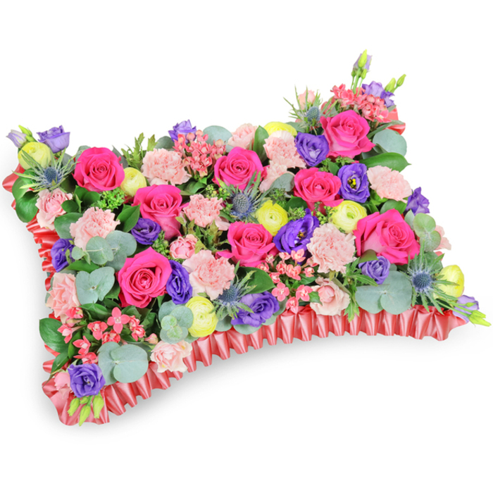 A rectangular pillow-style memorial arrangement is shown from a slightly elevated, angled perspective, the composition set against a pure white background to emphasise colour and form. The surface is a luxuriant mosaic of hot pink roses whose velvety blooms create the principal focus, softened by sprays of blush pink carnations that add delicate frill and volume. Bright yellow ranunculus appear like suns across the bed of flowers, and deeper purple lisianthus bring a cool, elegant contrast. Architectural hints of silver-blue eryngium thistle give an intriguing spiky texture, breaking the softness with a noble, wild note. Sprays of tiny pale filler blooms and generous swathes of rounded eucalyptus and mixed greenery cradle each bloom, providing depth, movement and a soothing, herbal scent. The entire cushion rests within a pleated, ruffled red satin border that frames the arrangement and reinforces the classic pillow silhouette; the ribbon's sheen catches the light and lends a refined finish. The lighting is gentle and natural, catching the curves of petals and the subtle shadows between layers of florals, conveying a tender, comforting atmosphere. Intended as a sympathetic floral tribute, this pillow communicates remembrance and celebration of life-an arrangement that would read beautifully at a service, placed at a chapel near Finsbury Square or at a family graveside, offering a warm, composed expression of enduring love and memory.