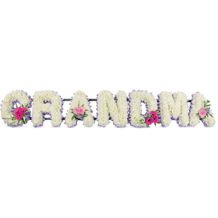 This photograph shows a thoughtfully arranged funeral tribute spelling the word GRANDMA in a line of bold, upright letters. Each letter is completely covered in pure white chrysanthemums, their small, many-petalled heads tightly packed to form a velvety, cushion-like surface. The flowers give the impression of softness and purity, with no gaps or stems visible, just waves of white petals flowing seamlessly across the shape of the word. Outlining each letter, a delicate band of lilac ribbon has been carefully pleated and attached, tracing every curve and corner with a subtle sheen. The ribbon introduces a gentle hint of colour that feels calm, respectful, and slightly nostalgic, as though chosen to echo a favourite cardigan or keepsake. Three focal posies interrupt the white surface at the first G, the middle A, and the final A. These clusters combine bright pink gerberas with wide, daisy-like faces, softer pink roses beginning to unfurl their central spirals, and sprigs of lush green foliage that add texture and depth. The greenery spills slightly beyond the edges of the letters, giving a natural, garden-inspired feel that would sit beautifully at a church or crematorium around Finsbury, perhaps after a quiet walk through Spa Fields or Myddelton Square Gardens. The lighting is even and gentle, bright enough to reveal each detail yet soft enough to maintain a sense of calm stillness. Though the image is silent, you can imagine the cool smoothness of the ribbon under your fingers and the fragile, almost silky touch of each chrysanthemum petal. This tribute carries the weight of memory and gratitude, expressing love for a grandmother through careful composition and the quiet language of flowers.