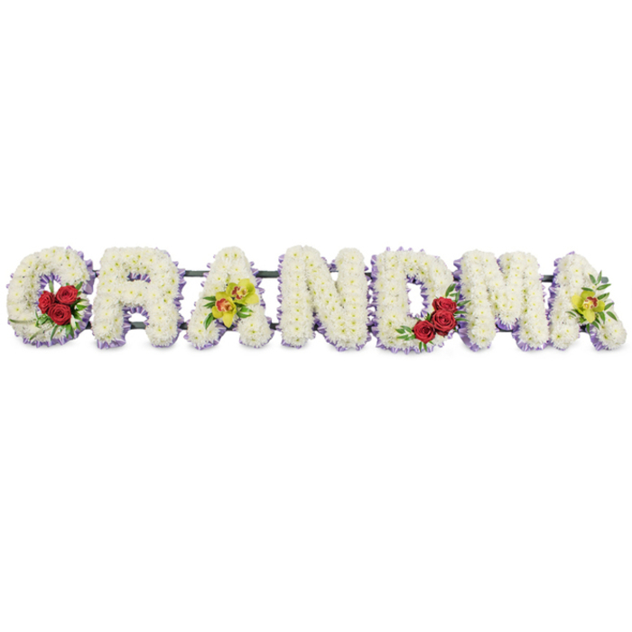 A refined, custom funeral flower arrangement in the shape of the word GRANDMA is the central subject of this image, laid out in a straight, unbroken line against a bright white background that emphasises both form and colour. Each uppercase letter is meticulously sculpted from a blanket of tightly clustered white chrysanthemums, their many small petals creating a soft yet structured surface, almost like a quilt of blossoms. Around the edges and along the base, a narrow underlayer of pale lavender flowers is visible, lending each letter a gentle lilac outline that adds dimension and a subtle hint of colour. On the opening G, three deep red roses are gathered into the lower curve, the blooms full and velvety, accompanied by well-groomed dark green foliage that provides natural contrast. The first A displays two striking chartreuse cymbidium orchids, carefully placed so their yellow-green petals and patterned centers rest on the diagonal bar, introducing a modern, slightly exotic feel amidst the classic white. The letters R, N, and D are left pure in white and lavender, their simplicity reflecting dignity and peace. The M features another trio of rich red roses tucked into its lower right, mirroring the placement on the G and drawing the eye along the word. The final A again carries two luminous yellow-green orchids, visually linking back to the first and closing the composition with gentle symmetry. A slender, dark metal frame runs behind the letters, providing strong but unobtrusive support for placement during a funeral or memorial. This thoughtful tribute could easily be imagined resting in a chapel near Finsbury, perhaps on a service route from Angel or King's Cross, offering a graceful, floral expression of love and gratitude for a grandmother who held the family together.