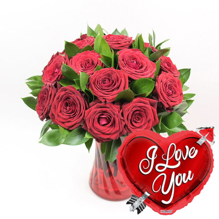 A richly romantic still life is shown against a pure white background: a dome of deep red roses rising from a ruby glass vase, accompanied by a bold heart-shaped balloon that seems to echo the same message in a playful tone. At the centre of the composition, about a dozen or more crimson roses are clustered tightly together, forming a rounded bouquet. Their petals are dense and velvety, layered in spirals that move from rich shadowed centres to softer, gently ruffled edges. Some flowers appear fully open, displaying broad faces of colour, while others remain slightly closed, adding variation and giving the bouquet a sense of life and unfolding. Among the blooms, fresh dark green foliage appears, leaves firm and glossy, giving structure and contrast, reminiscent of the greenery that lines quiet corners of Finsbury's residential streets. The stems collect in a translucent ruby-red glass vase, its fluted sides introducing vertical texture that catches the light in slender highlights. The water inside takes on a subtle red cast from the glass, adding depth to the base. In front of this arrangement, positioned in the lower right and overlapping the vase, a metallic red heart-shaped foil balloon adds a light-hearted flourish. The balloon's surface is highly reflective, showing bright white highlights against its saturated red colour. Across the middle, the phrase I Love You is written in smoothly flowing white cursive script, clear and inviting. A sleek silver arrow, an allusion to Cupid, appears to pierce through the heart diagonally, tying the design to the theme of romance. With no surrounding clutter, the clean white background allows the reds, greens, and metallic accents to stand out sharply, making this image perfectly suited to a Valentine's surprise near Finsbury, an anniversary gesture for a home by Amwell Street, or any occasion where you want the emotion of love to be unmistakable.