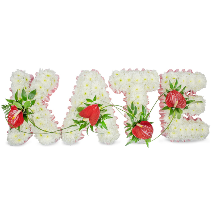 A carefully composed and dignified floral name tribute reads clearly as KATE in large, sculpted letters, each letter densely covered in small, delicate white spray chrysanthemums that give a uniformly soft, almost velvety surface suggestive of peace and purity. Around the outer edges a subtle, light-pink ribbon trim provides gentle colour and crisp definition, the pleated fabric catching light to outline each curve and angle without distracting from the florals themselves. Scattered through the white chrysanthemum field are clusters of glossy red anthurium blooms, their heart-shaped spathes and prominent yellowish spadices offering bold visual punctuation and warmth within the cool palette. Rich dark-green foliage-broad glossy leaves and slender fern fronds-has been artfully woven around the letters; fine green vines drift between the A and T, linking the individual forms and lending an organic continuity to the layout. Presented on a bright white backdrop with a faint drop shadow beneath the letters, the piece reads as three-dimensional and tactile, designed to be placed on a memorial table or standing easel so relatives can appreciate the craftsmanship up close. The overall impression is one of respectful elegance and thoughtful balance, a bespoke funeral tribute that combines classic sympathy flowers with refined florist technique. As a Finsbury-based design you might see near Finsbury Square or Exmouth Market, it conveys warmth, remembrance and the quiet dignity appropriate to commemorative services.
