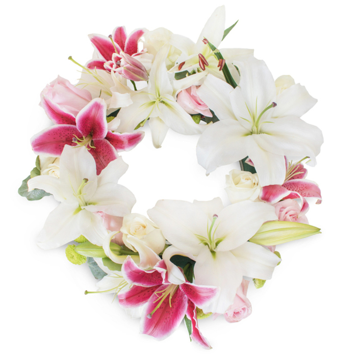 Standing out in luminous detail against a pure white field, this hand-arranged memorial wreath from Finsbury Flowers showcases the artistry and authenticity of a local florist. Large white Oriental lilies, their petals unfurling in abundant, star-like shapes, dominate the arrangement, some open wide to reveal soft green pistils, others just beginning to bud-each symbol of peace and remembrance the hallmark of thoughtful tribute. Accenting the whites, bold fuchsia and deep magenta lilies bring drama and vibrancy, enhanced by their subtle white speckles and the intensity of their colour, much like the vibrant street art or lively flower beds dotting Finsbury's many green spaces. Nestled gracefully between these prominent lilies, creamy and blush roses provide a romantic, velvety softness, their various stages of bloom adding the nuanced beauty of lives unfolding and remembered. Eucalyptus leaves and muted fresh greenery peek from behind, grounding the wreath with a touch of local nature and familiar scents of nearby parks. Every flower and foliage detail has been carefully selected, forming an unbroken ring that evokes unity, eternity, and enduring love-an ideal expression for commemorating a life well-lived, and for sending comfort across the close-knit Finsbury community.