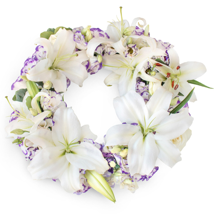 Photographed against an immaculate white background, this circular funeral wreath radiates a quiet, dignified beauty, as composed and reflective as a still moment in a Finsbury side street just off Rosebery Avenue. The design centres on an abundance of white Oriental lilies, each bloom a study in elegance with broad, velvety petals that curve gracefully away from a soft green core. The stamens rise slender and poised, their golden-brown anthers offering a delicate contrast to the cool white petals. Flowers at different stages of opening-some wide and star-like, others just beginning to unfurl from long, tapered buds-create a natural rhythm and a sense of life in motion. Threaded through these impressive blooms are clusters of Lisianthus, their petals softly frilled and tinged at the edges with shades of lilac, lavender, and muted violet. These smaller flowers fill the spaces between the lilies, lending the wreath a plush, gentle texture, like layers of folded fabric. Tiny creamy buds, likely roses or younger lilies, peek from the depths of the arrangement, adding nuance and the suggestion of tender, newly formed memories. A base of discreet greenery-slender dark leaves and fine eucalyptus-like sprigs in muted green-provides structure while maintaining the wreath's light, airy feel. The shape is a flawless circle, symbolising eternity, devotion, and the unending connection between the departed and those who remain. Created with the sensitivity of an experienced Finsbury florist, this wreath feels ideal for a memorial at a local church, a chapel near King's Cross, or a gathering to honour a life lived around Islington and Clerkenwell. Its restrained colours, soft textures, and balanced form offer a comforting visual solace at a time of loss.