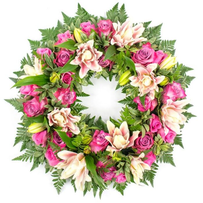 This image shows a beautifully crafted circular wreath set against a spotless white background, its colours and textures rendered with clarity that recalls a carefully arranged tribute in a Finsbury chapel or community hall. The wreath is dense and full, with flowers and foliage arranged so closely that they form a continuous, unbroken ring. Rich pink roses are the focal point, appearing in a range of shades from intense fuchsia and deep magenta to gentle blush. Some roses are tightly furled, their petals wrapped close in neat spirals, while others are fully unfurled with ruffled, velvety layers that soften the overall look and suggest openness and remembrance. Interlaced among these roses are elegant double lilies, whose broad, curling petals create a soft contrast in creamy white, pale pink, and the lightest peach. A few petals bear subtle deeper pink accents near the centre, while some lilies stand only partially open, with fresh lime-green buds hinting at blooms yet to unfold. The floral ring is bordered and supported by a lush foundation of greenery. Feathery fern fronds arc gently along the outer edge, their fine leaflets giving a light, airy texture, while robust, glossy dark green leaves anchor the design, filling empty spaces and adding visual depth much like the layered planting in Finsbury's pocket parks. The interplay of cool white background, warm pink tones, and deep green foliage creates a balanced, harmonious composition that feels both gentle and dignified. Though no ribbons or text are visible, the arrangement clearly reads as a sympathy wreath, suitable for funerals, memorials, and celebrations of life across Finsbury, offering a graceful floral symbol of enduring love, reflection, and respect.