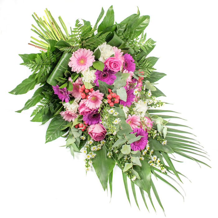 This asymmetrical funeral spray is photographed on a stark white surface, allowing its colours and structure to be clearly admired. The design flows in a diagonal from the neatly bundled stems at the upper left down to a tapering trail of foliage at the lower right, creating a dynamic yet serene line. Large gerbera daisies form the visual anchor of the piece, blooming in pale pink, vivid fuchsia, and deep magenta. Their wide, daisy-like heads add a sense of openness and optimism, balancing the somber occasion. Between them, classic pink roses with tightly spiralled centres emerge, their layered petals suggesting intimacy and love. Fluffy white blooms-likely small carnations or dianthus-are dotted throughout the composition, bringing a soft, ruffled contrast that brightens the palette. Slender, white, daisy-like fillers, reminiscent of chamomile, wander lightly among the larger flowers, introducing an airy, garden-inspired touch that softens the overall profile. Small clusters of bright red hypericum berries are nestled amid the petals and leaves, providing glossy, rounded accents that offer both richness and texture. The flowers rest on a dense foundation of greenery: broad, dark green leaves give structure and depth; fine, feathery fern fronds add movement and a slightly wild edge; and rounded, silvery eucalyptus leaves offer a cool, calming tone and the hint of a refreshing fragrance. At the lower right, long palm fronds stretch outward, reinforcing the spray's elongated silhouette. The effect is one of carefully orchestrated colour and texture, the sort of dignified tribute a family might order from Finsbury Flowers for a memorial near Sadler's Wells or around Old Street, hoping to express both sorrow and gratitude through a beautifully crafted floral farewell.