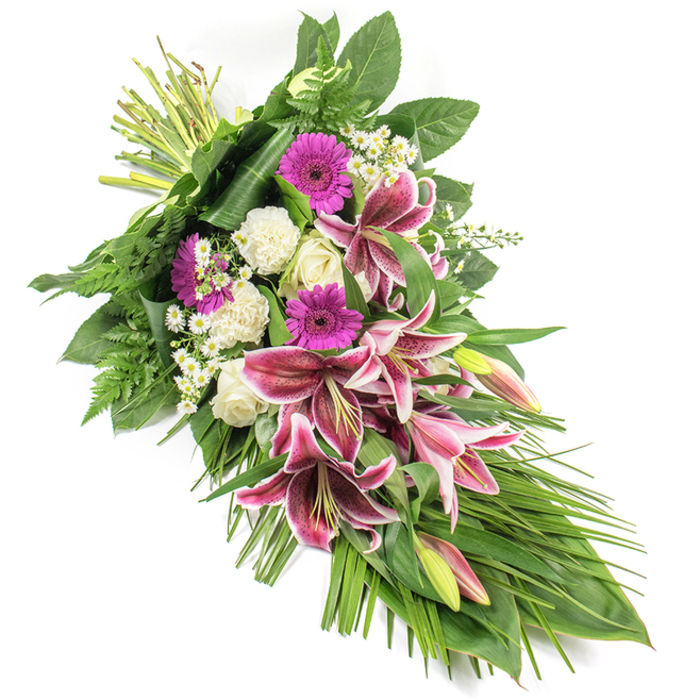 Set on a bright white background, this beautifully composed funeral spray extends in a long diagonal, from neatly bound stems at the upper left down to a soft, leafy cascade at the lower right, creating a teardrop silhouette that feels both flowing and composed-much like the blend of old and new streets around Finsbury. At the heart of the arrangement are several striking Stargazer lilies, each bloom painted in vibrant fuchsia-pink, framed by crisp white edges and dusted with burgundy speckles. Some blossoms are fully open, revealing their prominent stamens, while others remain in tall, elegant buds with hints of green and blush pink. Interspersed with the lilies are bold magenta gerbera daisies, their flat, round faces adding modern flair and an expression of fond remembrance. Soft, creamy white roses sit nestled among the brighter blooms, many still tightly furled, alongside gently ruffled white carnations that bring a soothing, cloud-like softness. Tiny white daisy-style fillers thread through the arrangement, bringing lightness, movement, and delicate detail. The flowers are held within a lush cradle of greenery: broad, dark green leaves form a strong structural base, while feathery fern fronds introduce texture and airy edges. Long, slender, blade-like leaves arc and trail downwards, accentuating the elongated form and guiding the viewer's gaze. At the top, the stems are carefully gathered and securely bound, evidence of expert, hand-crafted floristry often associated with local work prepared for services and family gatherings around Finsbury and nearby Angel. The spray's palette of rich pinks, calm whites, and layered greens creates a dignified, heartfelt tribute, ideal for accompanying a casket, standing beside a memorial, or conveying condolences when delivered to a bereaved home.