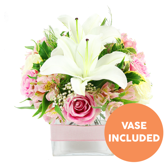 A beautifully balanced bouquet sits in a clear, square glass vase against a crisp white background, every element meticulously placed to highlight its form and colour. Dominating the centre are two large white Oriental lilies, fully open and slightly tilted, their silky petals stretching outward like soft trumpets and revealing pronounced reddish-brown stamens that add a dramatic focal point. Around these lilies cluster soft pink roses, their tones ranging from pale blush to a slightly deeper, warm pink, each rose at a different stage of opening: some neat and bud-like, others fully open with layered, velvety petals that suggest fullness and romance. Woven delicately between the roses are stems of pink and white alstroemeria, their slim petals etched with fine yellow streaks, adding an almost painterly detail and a sense of movement that feels right at home in the creative streets surrounding Finsbury. Fine sprays of white baby's breath float like tiny stars throughout the bouquet, lending a cloud-like softness and a light, airy feel. A rich collar of greenery frames the flowers, combining broad, glossy leaves with more feathery, fern-like fronds that give texture, depth, and a feeling of natural abundance. The square glass vase holding the stems is simple and modern, its middle encircled by a pale pink satin ribbon that introduces a gentle, decorative note, as if the bouquet were already wrapped for gifting by a local florist. In the bottom right corner of the image, a circular orange badge with bold white capital letters clearly reads VASE INCLUDED, signalling that the container is part of the arrangement. Overall, the composition conveys a sense of fresh elegance, ideal for marking special moments in Finsbury apartments, Highbury homes, or offices around Old Street.