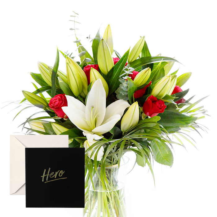 From the heart of Finsbury, there is joy in blending the bold with the serene, and this arrangement is a testament to that harmony. The white lilies drift at the nucleus of the bouquet, luminous and airy, their satin petals catching the daylight as if polished by a morning walk along Exmouth Market. Surrounding them, lush green foliage-dense, glossy leaves, slender ferns, and curling tendrils-forms a cool, verdant frame that makes the flowers glow. The red roses punctuate the design with a deep, velvet warmth, each bloom a quiet expression of respect and admiration. Unopened buds hint at ongoing beauty, inviting the eye to linger and anticipate what comes next. In soft, natural light, the arrangement feels buoyant and alive, a gift that seems to promise both strength and celebration. The texture of the petals-from the lily's satiny smoothness to the roses' velvety richness-speaks of careful handling and expertise. A gentle scent lingers-the clean sweetness of lilies, the subtle spice from the roses, and a hint of eucalyptus in the air. This hand-tied composition, with its balanced gravity and lift, is ideal for honoring someone's achievement or marking a milestone with quiet pride. A card marked 'Hero' accompanies the bouquet, making the gesture intimate and enduring, rooted in the care of your local florist in this corner of London.