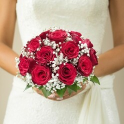 Amid the graceful architecture and historic charm of Finsbury, a bride holds a bouquet that radiates heartfelt tradition and refined elegance. The main feature-a cluster of about a dozen deep red roses-shows extraordinary richness, their tightly furled petals arranged in a rounded, opulent design. Laced amongst the roses, sprays of white gypsophila add a light, airy counterpart, their gentle blossoms almost hovering in contrast to the bold crimson blooms. Carefully placed vibrant green leaves offer a touch of woodland freshness, evoking the quiet gardens that dot Finsbury. The bride's hands, well-manicured and composed, tenderly cradle the bouquet, which rests above a gown of immaculate white. The gown reveals textured details, perhaps echoing the historic motifs found throughout the neighbourhood. A soft sash cascades softly to one side, reinforcing the sense of movement and anticipation. Subdued, creamy lighting ensures the bouquet's vivid colour and careful design are the clear visual focus. Celebratory yet composed, this arrangement is emblematic of Finsbury Flowers' artistry-perfect for those seeking bespoke wedding florals and the classic beauty of roses in the heart of London.