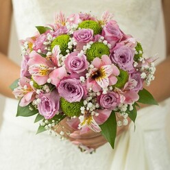 Amidst the urban charm of Finsbury, a bride's hands gently cradle a lush bridal bouquet-roses in soft pink and ethereal lavender unfurling at the core, their floral fragrance mingling with the excitement of a wedding day on Amwell Street. The bouquet is composed with an artisan's touch: vibrant button chrysanthemums in rich lime green provide a cheerful, contemporary accent and tactile contrast to the smooth rose petals, echoing the energy of local pocket parks. Pale pink alstroemeria lilies, delicately marked with maroon and gold streaks, peek out from the blossoms, imparting a sense of refinement and an occurrence of whispered grace, reminiscent of art exhibitions just off Exmouth Market. Airy sprays of gypsophila float overhead, creating a soft, romantic halo that brings a touch of lightness and dream-like candour. Glossy, dark green foliage supports the arrangement, grounding the pastel palette. Made for those drawn to the blend of tradition and innovation that defines Finsbury, this bouquet sparkles with heartfelt artistry-a fitting centrepiece for a wedding near the historic squares, and a testament to the skill of local florists who capture the beauty of every love story and city celebration.