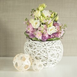 Bathed in soft natural light, this wedding table arrangement embodies Finsbury's blend of charm and contemporary elegance. The centrepiece features a spherical white vase, artfully crafted from open-weave rattan that channels the handmade aesthetic of nearby local shops. Rising from the vase's airy structure is an exuberant bouquet of creamy eustoma and pale yellow lisianthus, their delicately ruffled petals unfolding with the promise of spring mornings in Spa Fields gardens. Pink stock flowers rise elegantly amidst the lisianthus, introducing gentle height and an understatedly sweet scent that recalls flower stalls lining Chapel Market. Beneath these main blooms, vibrant purple fillers-suggestive of aster or allium-bring a sense of depth and local vibrancy, highlighted by encircling sprigs of glossy dark green foliage. Off to the left, two sculptural spheres fashioned from pale rattan echo the open-work design of the vase, evoking the creative spirit and tactile warmth of Finsbury's craft scene. The arrangement sits on a sleek white tabletop that catches the diffused daylight so characteristic of Finsbury's bright interiors. A subtle greige wall with faint vertical texturing in the background provides a calming, modern backdrop, elevating the gentle pastel hues. Infused with artistry and care, this arrangement is perfect for wedding celebrations and intimate gatherings, offering a taste of local creativity and Finsbury freshness, available for bespoke event delivery from the team at Finsbury Flowers.