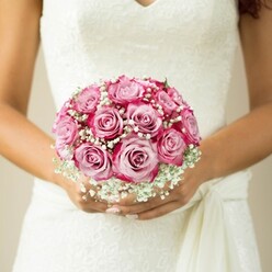 This bridal bouquet, beautifully hand-tied and radiating modern grace, feels right at home in the elegant ambiance of Finsbury. The arrangement showcases an array of two-tone roses-pale pink at their centre, deepening to a dramatic magenta at the rim-each bloom a delicate echo of passion and serenity. Billows of delicate white baby's breath entwine among the roses, imparting a dreamy, contemporary finish reminiscent of the boutique wedding scenes found on the edges of Finsbury's vibrant community spaces. The bride's gently bronzed hands, nails touched by a soft nude polish, cradle the flowers with affection and quiet anticipation. Her gown, all sleeveless white lace and finely sculpted lines, serves as the perfect backdrop, while a loosely tied sash detail peeks out just above her hands, nodding to modern bridal design seen at Finsbury Flowers. The bouquet is the undeniable focus, with the simple, beige-blurred background letting the colours and textures sing, capturing an air of tranquil excitement that so many Finsbury ceremonies embody. This scene is a heartfelt celebration of fresh floral design, inspiring local brides with its blend of classic romance and urban style.