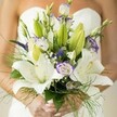A vision of delicate romance, this Finsbury bridal bouquet is alive with soft ivory lilies and swirls of purple-edged lisianthus, their forms opening in a slow, hopeful choreography. The creamy petals of the lilies gleam gently beneath the shy morning light, while unopened buds whisper of stories awaiting their turn-a graceful nod to the promise of new beginnings. Ruffled lisianthus petals, painted in watercolour shades of lavender, curl playfully among slender spikes of royal purple Veronica, the bouquet's lively energy recalling the vivacity of Finsbury's historic streets and leafy parks. Baby's breath forms airy halos around the lilies, and feathery sprays of asparagus fern lend movement and texture, as if the bouquet itself were caught in a light breeze drifting down Clerkenwell Road. The bride's softly manicured hands gather the stems, above which the blurred suggestion of a white, strapless gown completes the scene. The ensemble radiates purity, artistry, and joy-a heartful creation by Finsbury Flowers, fit for an urban celebration or a private moment in the gardens that grace this vibrant corner of London.
