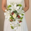 A bride stands against the softly blurred backdrop of a Finsbury wedding suite, holding an exquisite cascading bouquet that brims with artistry and local charm. Her hands, just visible above a sea of petals, support a generous arrangement where snow-white lilies fan outward in striking starbursts, their vibrant form accented by gentle spots of earthy brown. Nestled within is a tightly coiled rose in creamy pale yellow, bringing a classic romantic accent echoing the timeless style seen throughout Finsbury's historic passages. Anchoring the arrangement, clusters of lively lime-green flowers and plump red hypericum berries create a lively interplay of colour, their freshness evocative of early morning strolls through leafy Amwell Street. Wispy sprays of baby's breath lend a dreamy, cloud-like aura, gently framing each larger flower. Cascading from the bouquet's core, icy-white Phalaenopsis orchids trail gracefully downward, capturing the motion and elegance of a true teardrop or waterfall bouquet. Framing the display, rich green foliage and slender arches of grass conjure the feeling of wild meadows at the city's edge. A subtle satin ribbon ties the stems together near the bride's hands, adding a whisper of classic refinement to the contemporary palette. This bouquet, hand-picked for a Finsbury celebration, captures the balance of traditional beauty and fresh innovation - perfectly suited to weddings held in the heart of London's vibrant north.