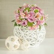 A serene and intricately handcrafted centerpiece takes center stage in this softly illuminated image, echoing the creative sensibilities of Finsbury's close-knit community. The main bouquet is a gentle cascade of full-bodied pastel pink and pale lavender roses, their velvety petals layered among the refined beauty of pink alstroemeria with their signature striped throats. Offsetting these classic blooms, fresh lime green button chrysanthemums tuck into the arrangement with bright cheer, while delicate sprigs of baby's breath dance in between, contributing to a sense of airy elegance that brings to mind peaceful strolls through Finsbury parks. The arrangement is housed in a striking white vase-its open lattice design, constructed from finely woven strips, recalls both contemporary style and classic basketry admired at the local artisan shops. On the left side of the vase, two decorative spheres in natural, light shades add harmonious detail; their irregularly woven forms provide visual continuity and playful sophistication. The entire ensemble is set against a clean white table, glowing under soft light, and framed by a textured beige backdrop that lets the floral colours and tactile elements stand out. Accented by rich green leaves, this centerpiece radiates warmth, elegance, and an understated celebration of Finsbury's artistic culture-ideal for wedding tables, creative events, or simply to bring joy and freshness to any room it graces.